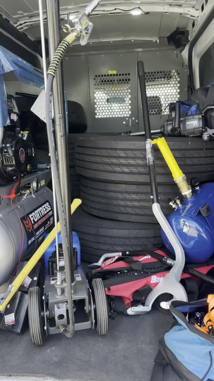 Inside the van: a gas compressor, a floor jack with a long handle, a stack of truck tires, a blue portable air tank, a tire iron, and a red strap bag