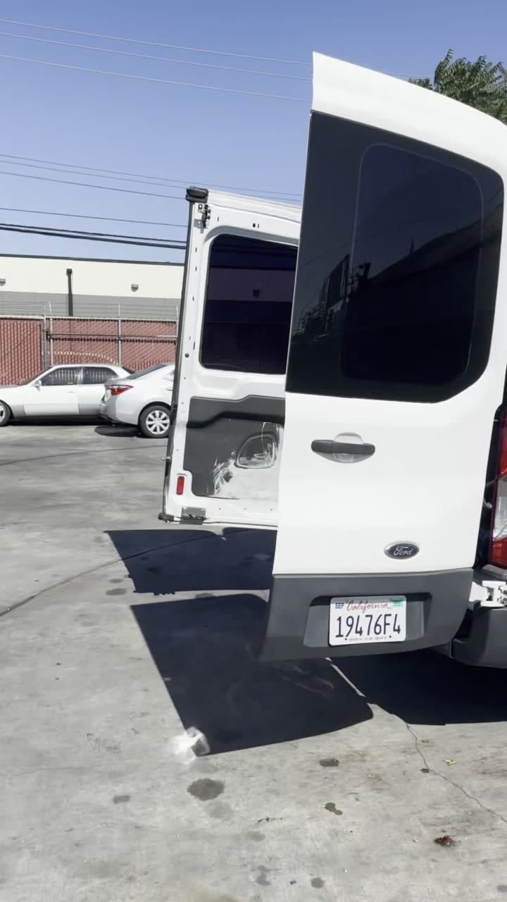 White service van seen from behind with one rear door swung open, parked on a paved lot beside a chain-link fence